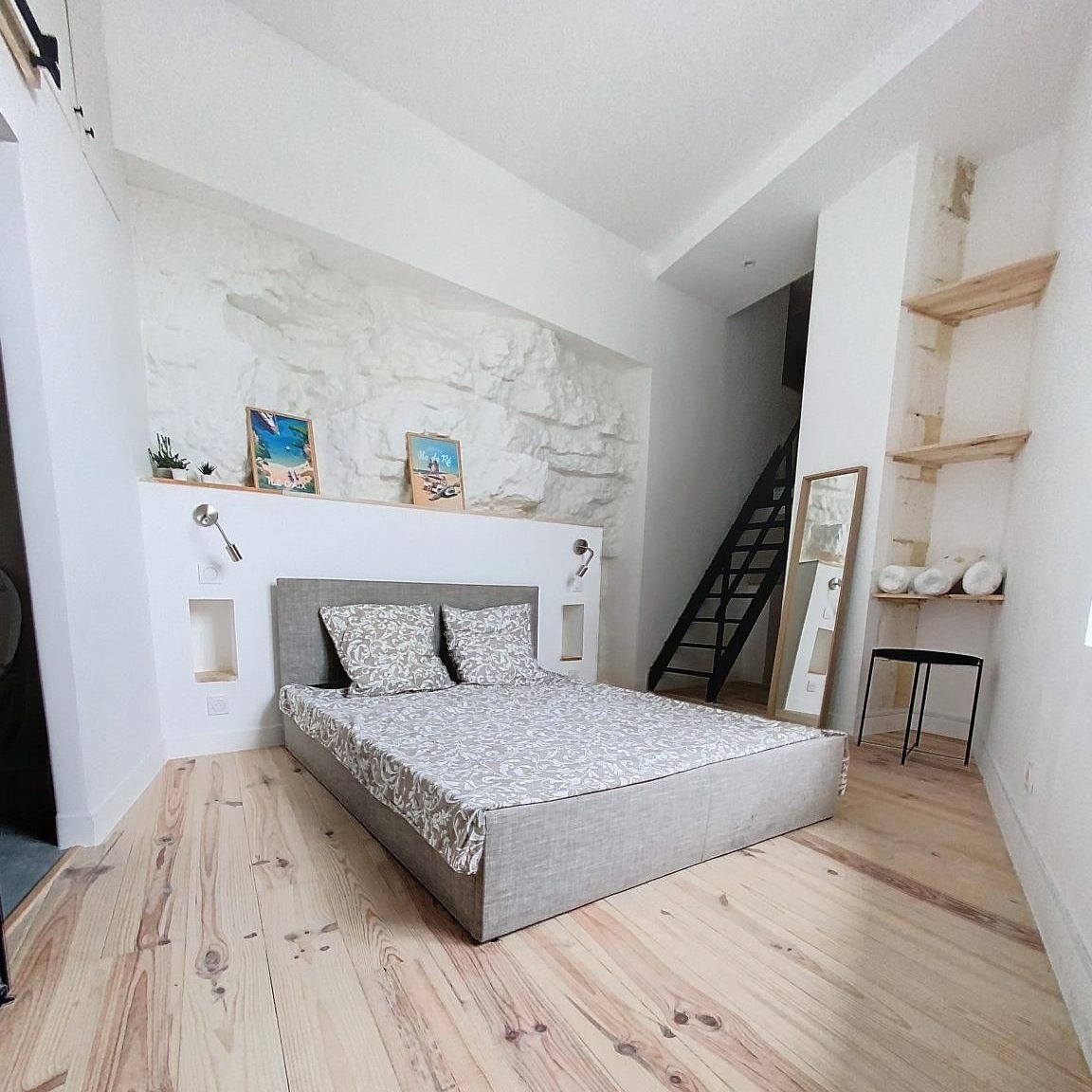 Chambre lumineuse avec un lit au sol et des murs en pierre blanche. Étagères et décor minimaliste.