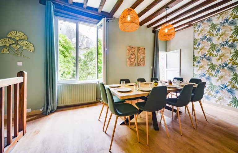 Comedor luminoso con mesa grande, sillas de colores y ventanas abiertas.