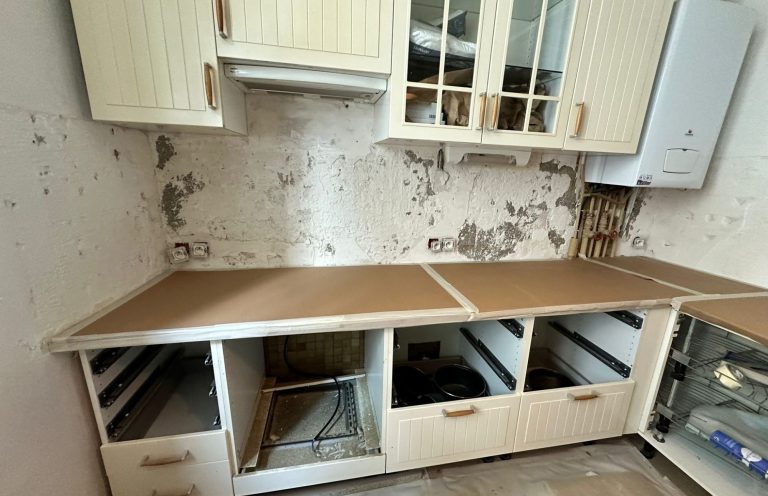Cocina con gabinetes de madera clara y paredes deterioradas. Encimeras vacías y desordenadas.
